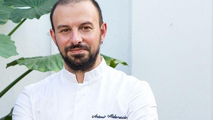 Bearded vegan chef Antonio Alderuccio smiling with a white chef top on with a house plant in the background