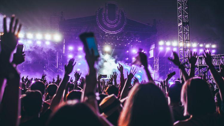 A crowd of people with their hands in the air at a music festival in the evening in front of a brightly-lit stage