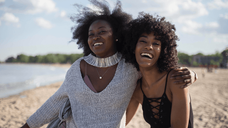 Woman besides another woman at the seashore both smiling and embracing one another